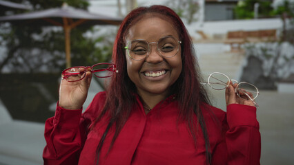 Woman wearing glasses smiles while holding red and clear eyeglass frames near face on street; fashion decision joy.