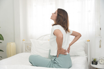 Young woman touching her lower back in pain while sitting on bed, showing signs of backache or...