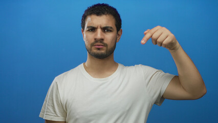Young hispanic man points finger down with frowning face in isolated blue studio with serious...