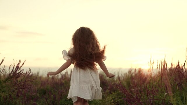 Happy little girl runs enjoying freedom in field at summer sunset backside view. Active little girl runs across rural field on summer vacation. Healthy little girl plays running across field at sunset