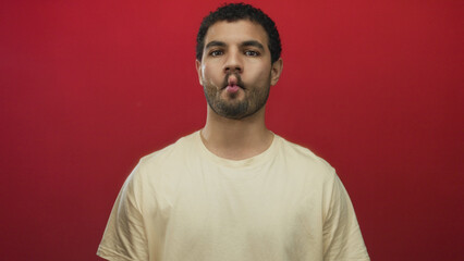 Fototapeta premium Young hispanic man with curly hair and beard stands and purses lips in a fish face pose in a red studio; joy.
