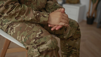 Fototapeta premium Young man in camouflage uniform sits on a chair and clasps hands inside a building; quiet reflection.