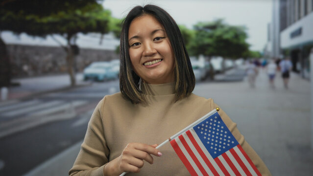 Young chinese woman holding american flag smiling on a city street, symbolizing cultural diversity and international connection in an urban outdoor setting.