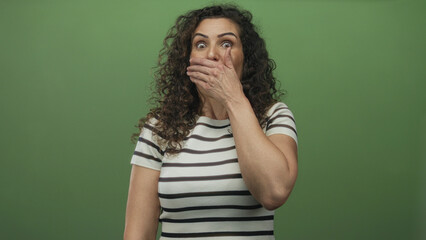 Young hispanic woman covers mouth with hand and points thumb to side in green studio; surprise reaction.