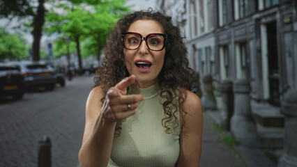 Woman points finger at camera on busy street lined with cars under leafy green trees in bright...