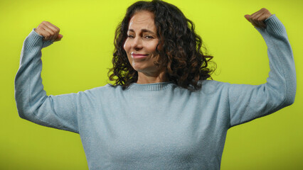Woman flexing confident arms in blue sweater against bright yellow background in studio shot.