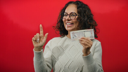 Woman smiling and pointing while holding uae dirham banknote against a bold red wall background,...