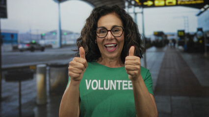Hispanic woman in volunteer uniform smiling with thumbs up at an airport terminal outdoors, conveying positivity and helpfulness in a busy travel environment.