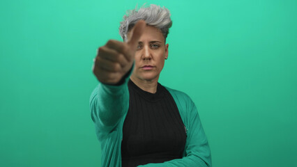 Woman with short hair shows thumbs up gesture in green studio setting, wearing teal cardigan and...