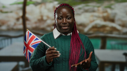 Woman holding british flag in outdoor terrace setting wearing green sweater with red braids, symbolizing multicultural identity in city backdrop emphasizing unity and diversity.