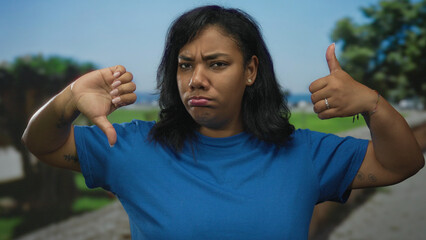 Woman wearing blue shirt shows thumbs up and thumbs down gesture in sunlit park with blurred trees;...