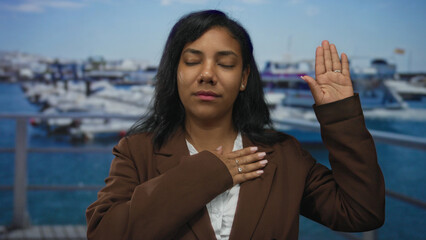 Woman in brown blazer raises hand and places palm on chest on street by waterfront under blue sky;...