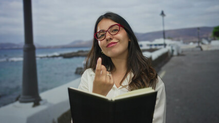 Fototapeta premium Young hispanic woman with red glasses reading black notebook on seaside promenade by gentle ocean waves; tranquility.