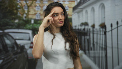 Fototapeta premium Hispanic woman salutes with two fingers at temple on street beside wrought iron fence and parked car; friendly greeting.