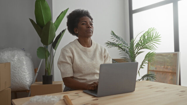 Woman working on laptop surrounded by moving boxes and plants in a new home living room suggesting a peaceful work from home environment.