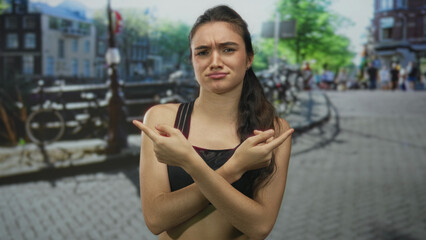 Young hispanic woman wearing a sports top points finger along a cobblestone street by a canal railing; confusion.