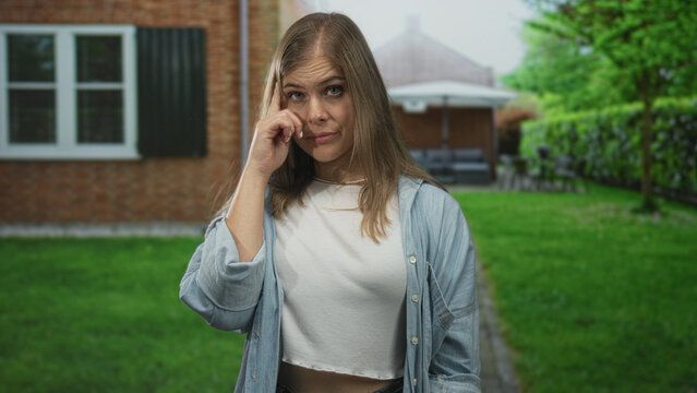 Woman in denim shirt points finger at camera in front of building entrance, standing on pathway; defiance.