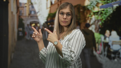 Blonde woman in glasses pointing finger down narrow cobblestone street beside outdoor cafe tables; uncertainty.