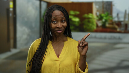 Woman wearing yellow shirt with curly hair gestures expressively on urban street background indicating outdoors scene.