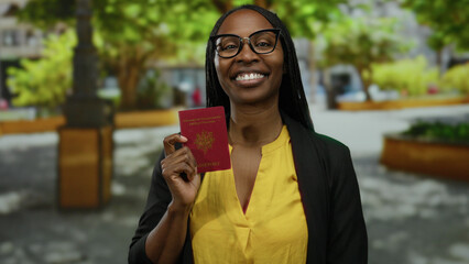 Woman holding french passport outdoor in park setting, smiling confidently, wearing glasses and...