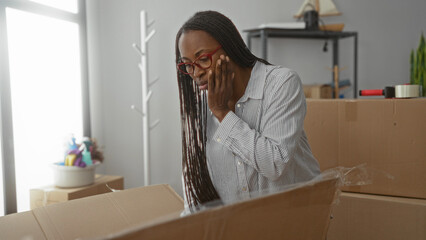 Woman unpacking box in new home with thoughtful expression, wearing glasses and striped shirt, surrounded by moving supplies in apartment living room.