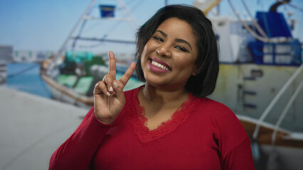 Woman smiling at port with boats in background displaying peace sign in red sweater on sunny seaside day showing joy and positivity outdoors.