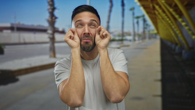 Young man outdoors in city making mock crying gesture with a playful expression against an urban street background, suggestive of humor and sarcasm in a vibrant urban environment.