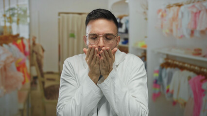 Naklejka premium Young hispanic man in a white coat feeling cold in a colorful clothing shop, hands pressed together, surrounded by racks filled with vibrant garments, creating a cozy shopping scene.