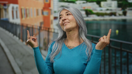 Woman with grey hair makes expressive gestures on seaside promenade, wearing blue sweater, surrounded by beach scenery, embodies outdoor freedom and defiance in vibrant setting.