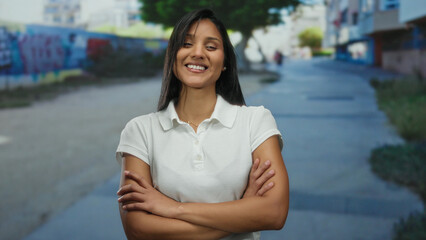 Woman smiling confidently on a vibrant urban street, arms crossed, featuring a colorful graffitied wall and bright daylight, dressed casually in a white shirt outdoors.