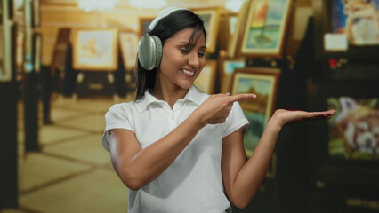 Woman smiling in art gallery wearing headphones making ok gesture, showcasing creativity and joy indoors with framed artwork in background, expressing positivity.