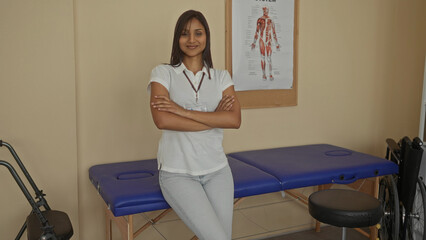 Young woman in clinic room smiling with arms crossed beside a treatment table, showcasing a professional medical environment with anatomical poster and wheelchair.
