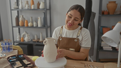 Young hispanic artisan woman holds ceramic vase with hands in studio surrounded by pottery shelves and sculpting tools; serenity.