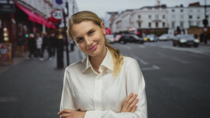 Fototapeta premium Woman smiling confidently in a city street with arms crossed wearing a white shirt outdoors in an urban european setting with traffic and pedestrians in the background.