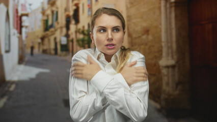 Obraz premium Woman in white shirt feeling cold in an old city street, embracing herself to stay warm amidst the medieval outdoor scenery.