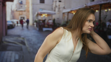 Woman holds hand to ear leaning forward and listening on a busy cobblestone street under evening light; confusion.