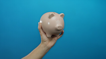 Hand holding a piggy bank against a solid blue background, illustrating financial savings and...