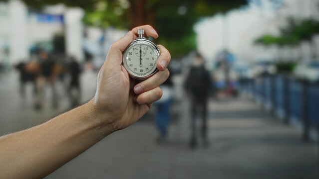 Caucasian man holding stopwatch on city street with blurred background showing urban outdoor environment, emphasizing timing and urgency in public spaces.