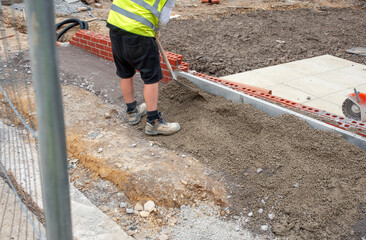 Builder working on sidewalk installation using shovel to place concrete around new edging pin kerb