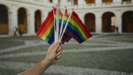 Hand holds vibrant lgbtq rainbow flags outside in a city street, symbolizing pride and solidarity, with historic architectural background and an outdoor setting.