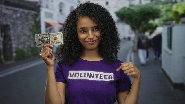 Woman holding dollar in street wearing volunteer shirt smiling outdoor young latin hispanic female showing american money