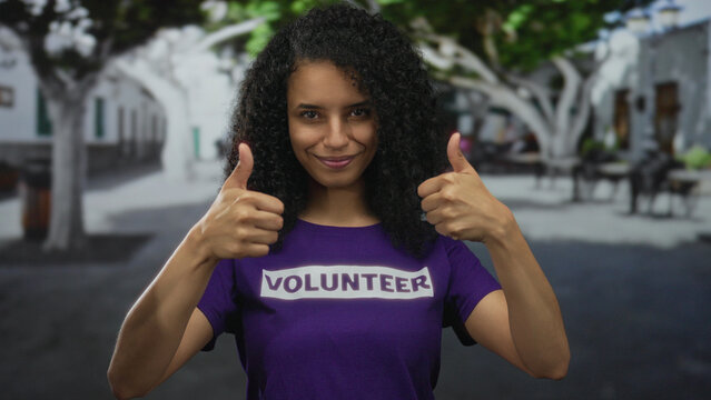 Woman with curly hair wearing volunteer shirt giving thumbs up on a street backdrop outdoors showcasing positivity and community spirit