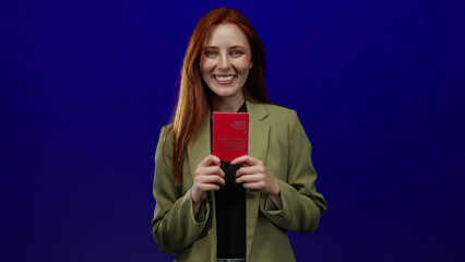 Woman holding swiss passport smiling in front of isolated blue background showcasing identification travel document adult redhead female attractive