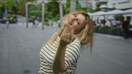 Woman beckons with finger while smiling on busy city street by sidewalk cafes and blurred...