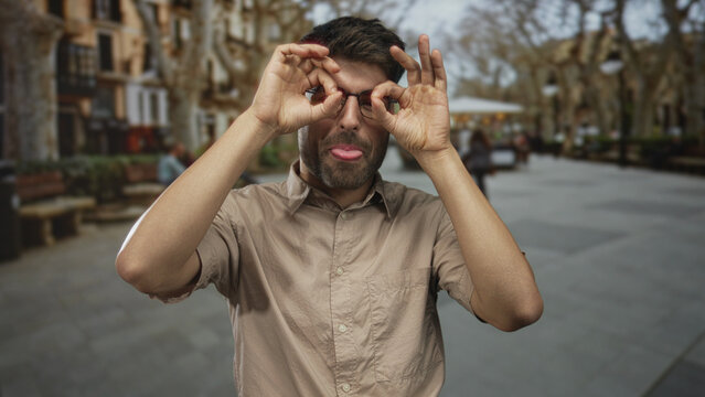 Young hispanic man frames eyes with both hands as binoculars on bustling city street plaza; curiosity.