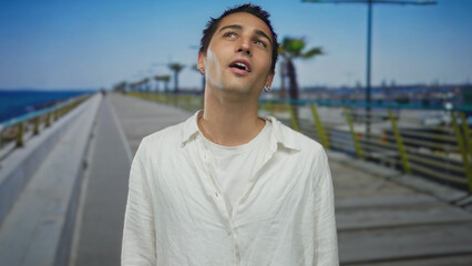 Young hispanic man in white shirt aims finger gun to his temple on a sunny seaside promenade; frustration.