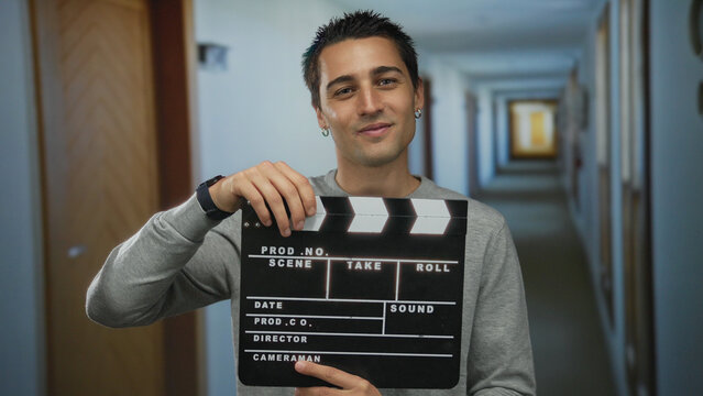 Young hispanic man holding clapperboard in hotel corridor creating film scene enthusiastically indoors representing filmmaking process and cinematic storytelling essence