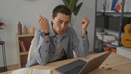 Young man in a home apartment sitting at a desk with a laptop, gesturing and smiling confidently in a modern living room setting.