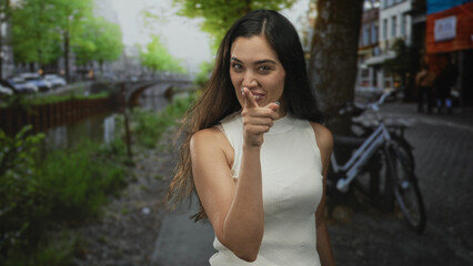 Woman smiling and pointing finger at camera on a busy canal street by the water with bicycle in view; confidence.