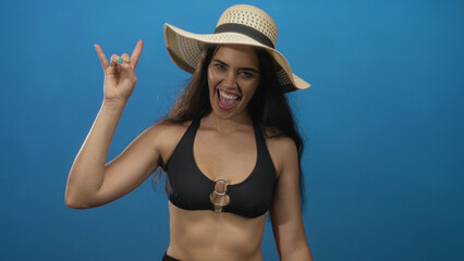 Woman in a blue studio wearing black bikini top and straw hat making rock horns gesture; playfulness.